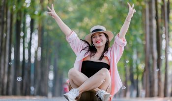 Smiling woman sitting cross-legged on a scenic tree-lined road, wearing a hat and raising peace signs.