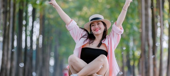 Smiling woman sitting cross-legged on a scenic tree-lined road, wearing a hat and raising peace signs.